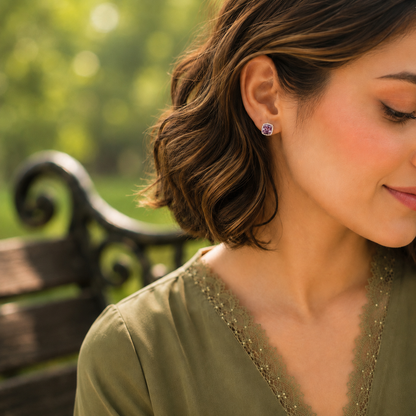 Close-up of a woman wearing silver halo stud earrings with a cushion-cut purple crystal in a soft outdoor garden setting.