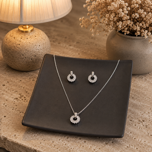 Silver halo crystal pendant and matching earrings set displayed on a charcoal grey ceramic plate over a premium travertine stone surface with a luxury blurred background featuring a lamp and dried flowers.