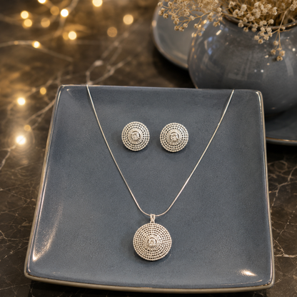Silver crystal disc pendant necklace with matching round stud earrings displayed on a smokey blue ceramic plate over a premium reflective marble surface with softly blurred fairy lights and a matching ceramic vase.