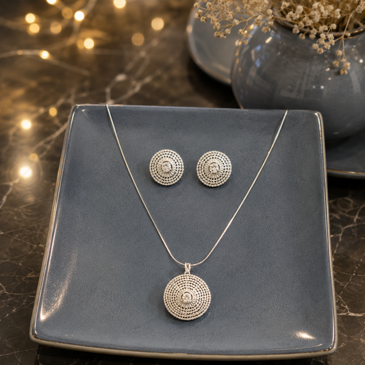Silver crystal disc pendant necklace with matching round stud earrings displayed on a smokey blue ceramic plate over a premium reflective marble surface with softly blurred fairy lights and a matching ceramic vase.