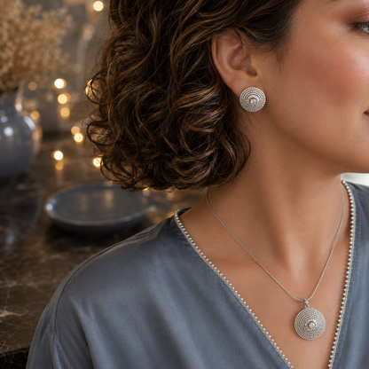 Close-up side profile of a model wearing a silver crystal disc pendant necklace and matching stud earrings with a smokey blue satin top featuring a pearl-trimmed band neckline in a luxury studio setting.