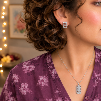 Close-up side profile of a model wearing a silver geometric rectangular pendant necklace and matching crystal stud earrings with a plum floral linen outfit in a luxury lifestyle setting.