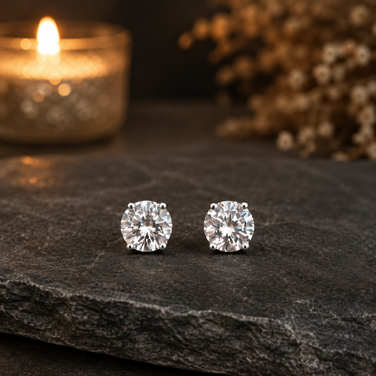 Classic round solitaire crystal stud earrings displayed on a dark grey stone surface with a softly blurred candle-lit background.