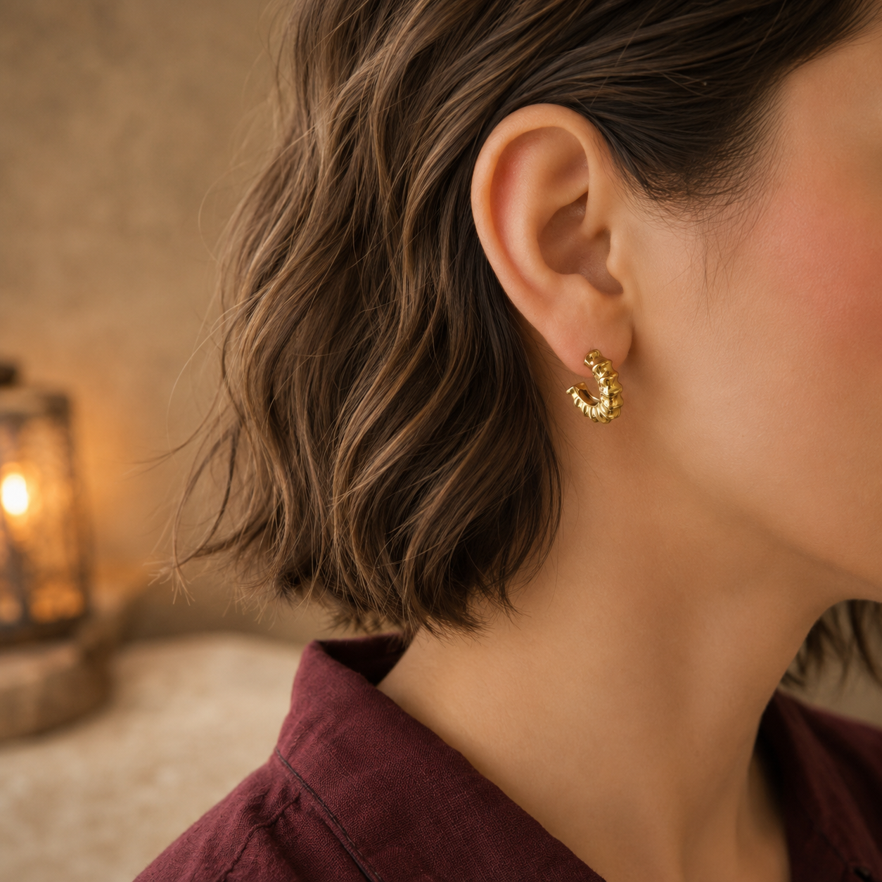 Woman wearing textured gold mini hoop earrings with a maroon linen shirt in a close-up portrait.