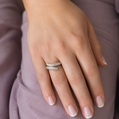 Woman wearing a silver twisted pavé wrap ring with sparkling micro-set stones on her hand, photographed in a luxury editorial style with a soft mauve background