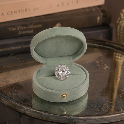 Silver oval halo statement ring with an oval-cut centre crystal, baguette halo and pavé halo displayed inside a premium sage green oval jewellery box on an oxidized glass t