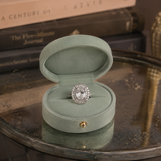 Silver oval halo statement ring with an oval-cut centre crystal, baguette halo and pavé halo displayed inside a premium sage green oval jewellery box on an oxidized glass t