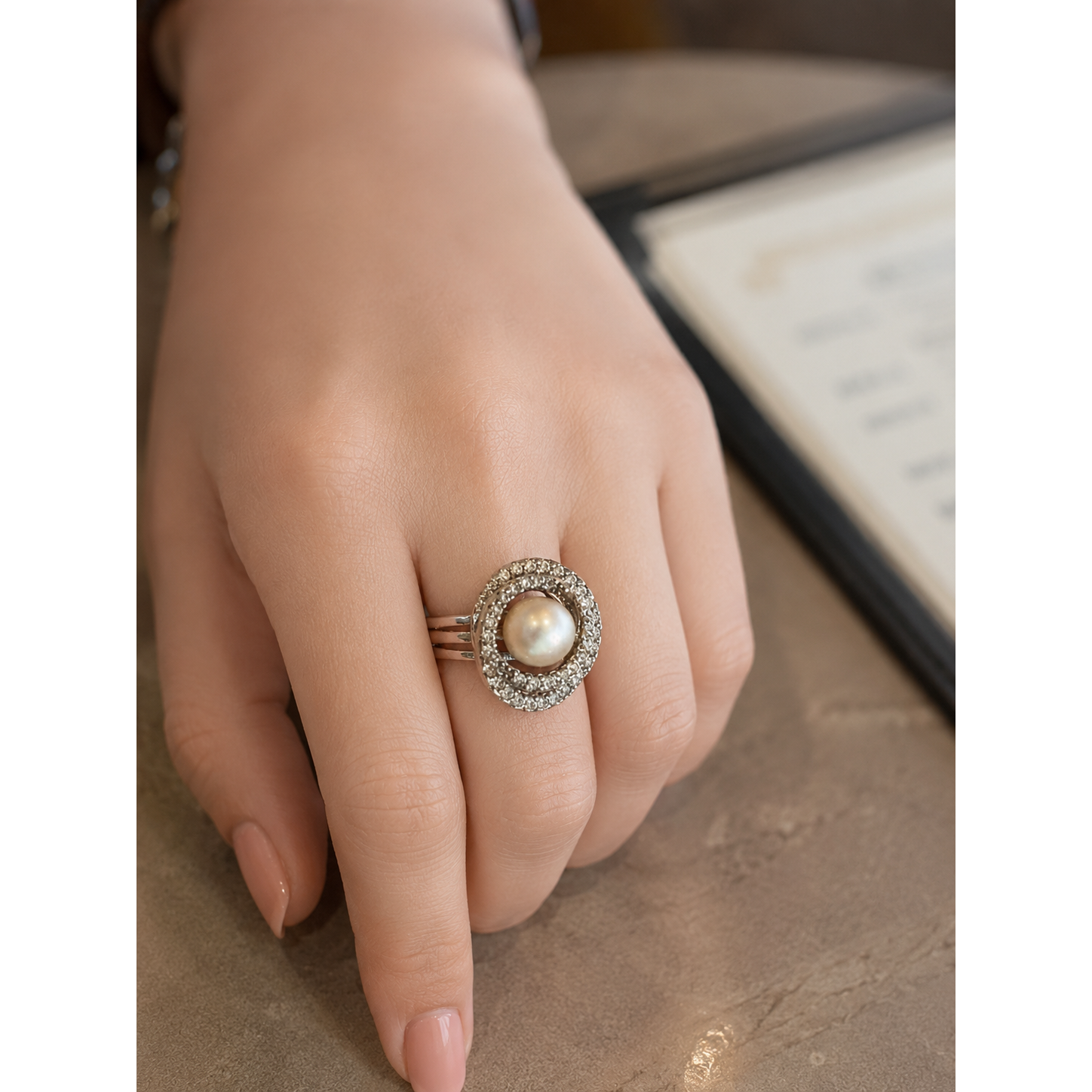 Woman wearing a polished silver statement ring with a round pearl centre encircled by shimmering zircon stones, photographed on a café table with a softly blurred menu in the background.