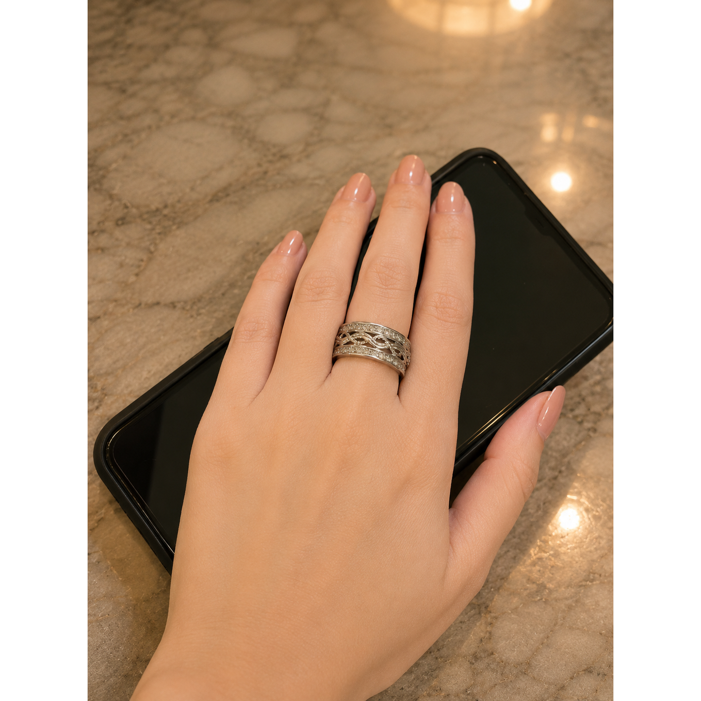 Woman wearing a wide silver infinity band with interwoven openwork detailing and sparkling zircon borders while resting her hand on a black mobile phone over reflected marble