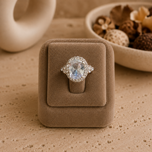 Silver-tone oval halo crystal ring displayed on a champagne premium ring stand over a travertine surface with softly blurred ceramic décor and potpourri.