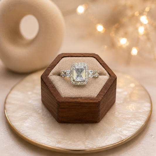 Silver halo ring with emerald-cut clear center stone displayed on a premium wooden ring stand over a pearlized tray with softly blurred ceramic décor and fairy lights.