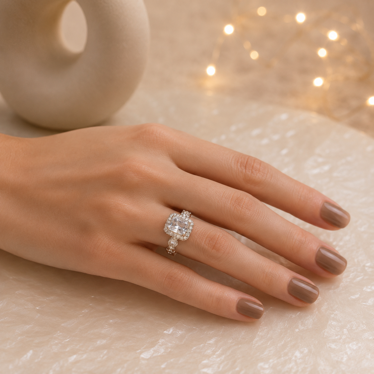 Woman wearing a silver halo ring featuring an emerald-cut clear center stone and marquise side accents on a smooth manicured hand against a pearlized luxury surface.