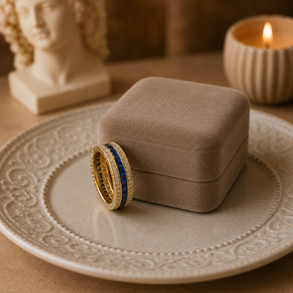 Gold eternity band with royal blue princess-cut gemstones leaning against a premium taupe velvet jewellery box on an embossed ceramic plate with warm ambient styling.