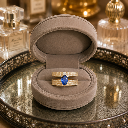 Light gold open-back double-band ring featuring a pear-cut sapphire blue centre stone with sparkling zircon detailing displayed in a luxury taupe velvet jewellery box.