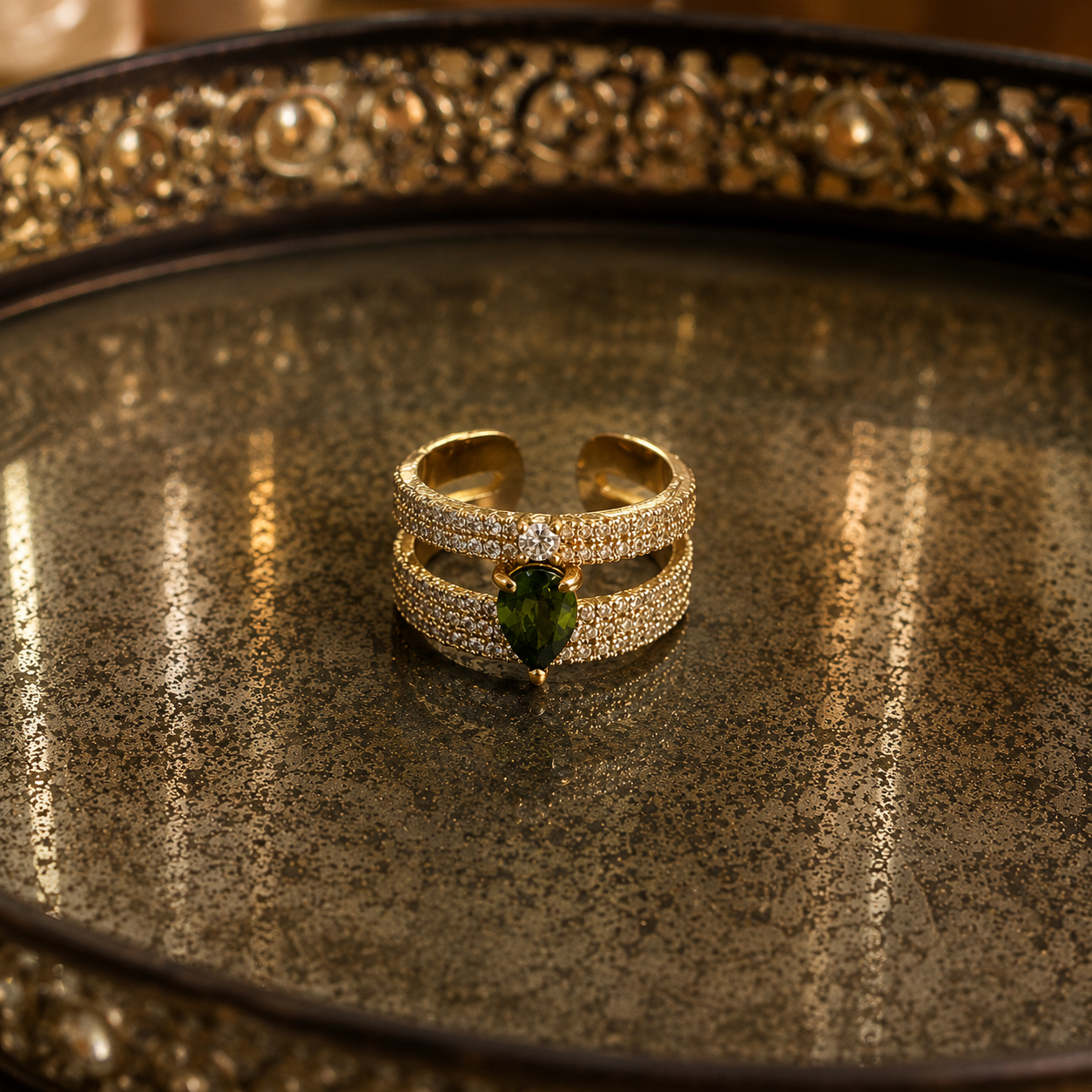 A gold-toned double-band ring with pavé-set clear stones and a central green marquise-cut gemstone, displayed on a textured metallic tray.