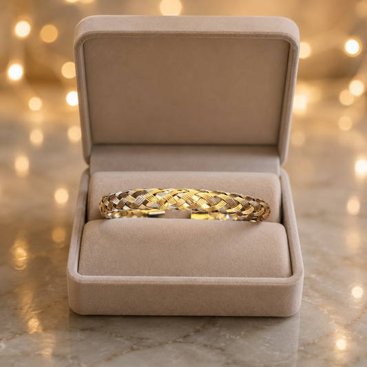 A gold braided cuff bracelet displayed inside an open beige velvet jewelry box on a marble surface with warm bokeh lights in the background.