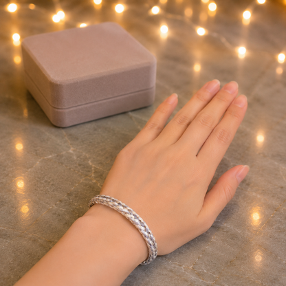 A silver braided bangle bracelet worn on a person's wrist, with a matching dusty pink jewelry box in the background on a gray marble surface decorated with warm string lights.