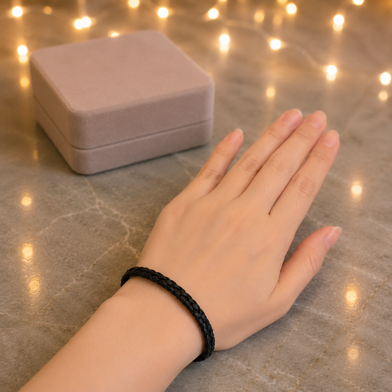 A black braided cord bracelet worn on a person's wrist, with a small dusty pink jewelry box in the background and warm string lights on a gray marble-like surface.