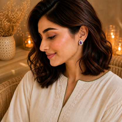 Woman wearing silver teardrop stud earrings with an amethyst purple pear-cut centre stone and pavé halo in a warm luxury indoor setting