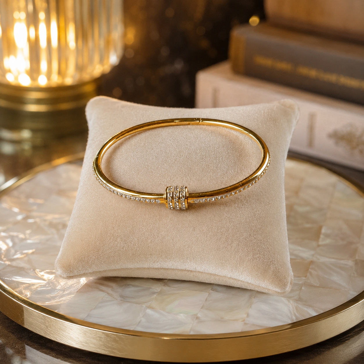 Polished gold crystal cuff bracelet with a secure twist-lock screw closure displayed on a cream velvet cushion atop a pearlized round tray with softly blurred books and warm ambient lighting.