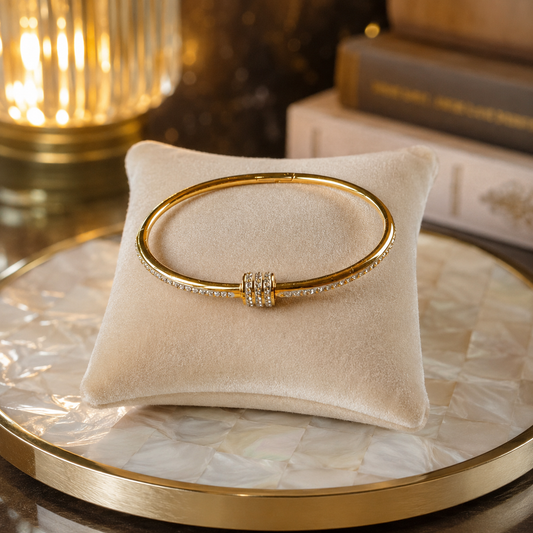 Polished gold crystal cuff bracelet with a secure twist-lock screw closure displayed on a cream velvet cushion atop a pearlized round tray with softly blurred books and warm ambient lighting.