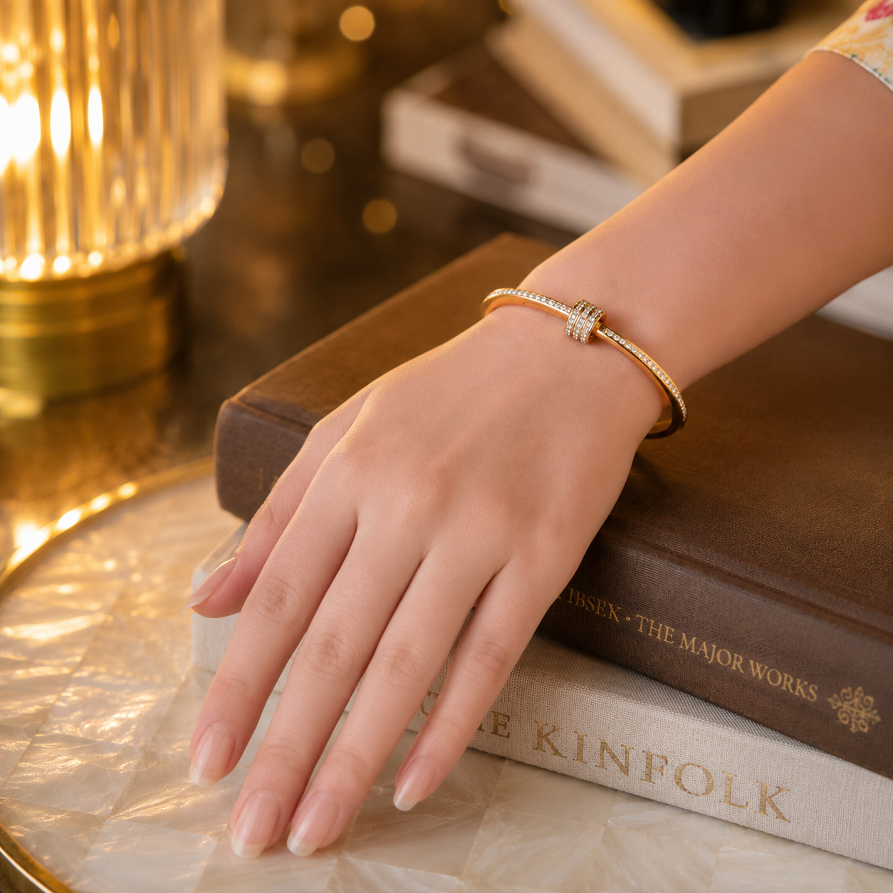 Elegant gold crystal twist-lock cuff bracelet worn on a feminine hand resting on stacked books with a warm ambient lamp and pearlized tray in the background.