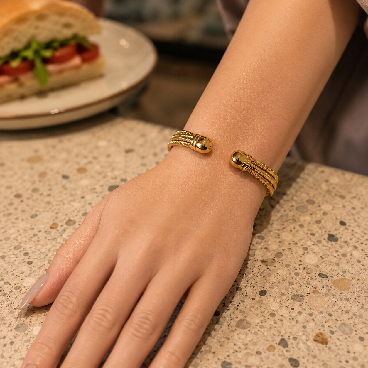 Premium gold multi-strand open cuff bracelet with polished rounded ends worn on a feminine wrist in a warm café lifestyle setting.