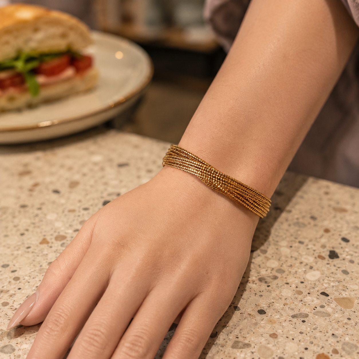 Elegant gold multi-strand twist cuff bracelet worn on a feminine hand with smooth skin and neutral-toned nails on a modern café countertop.