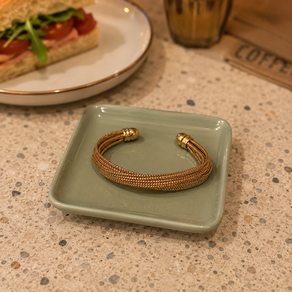 Luxury gold multi-strand twist cuff bracelet displayed on a premium sage green ceramic plate in a stylish café setting with a softly blurred sandwich in the background
