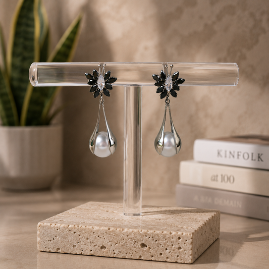 Luxury black crystal floral pearl drop earrings displayed on a clear acrylic stand with a travertine stone base in a premium editorial jewelry setti