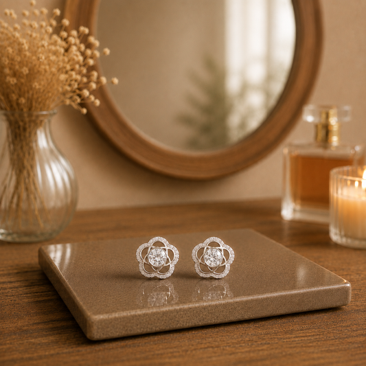 Silver floral stud earrings with clear crystal center stones displayed on a premium ceramic tile with blurred wooden mirror, perfume and dried flowers.