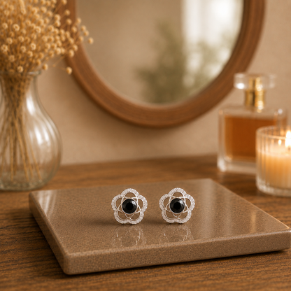 Silver floral stud earrings with black crystal center stones displayed on a premium ceramic tile with blurred wooden mirror, perfume and dried flowers.