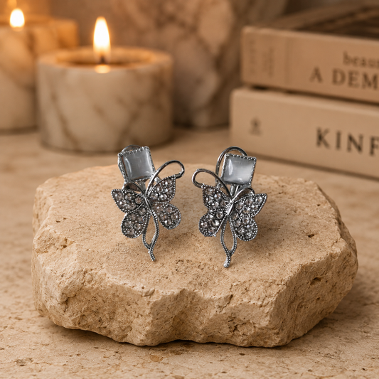 Silver butterfly crystal earrings displayed on a beige travertine stone pedestal with a luxury candle-lit background.