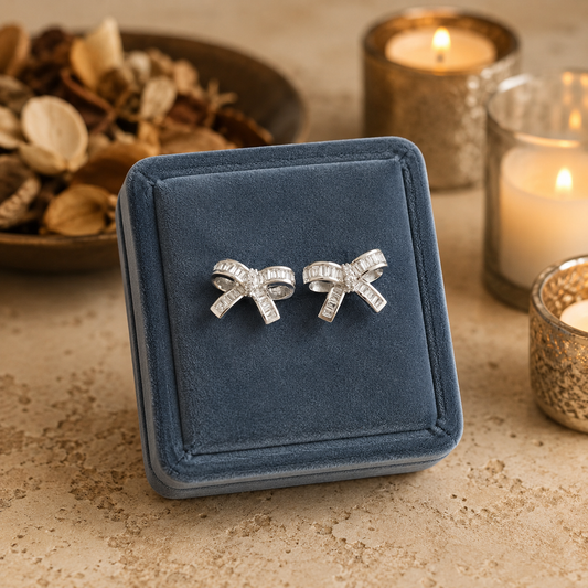 Silver crystal bow stud earrings displayed on a premium smokey blue velvet jewellery stand with softly blurred candles and potpourri on a textured limestone surface.