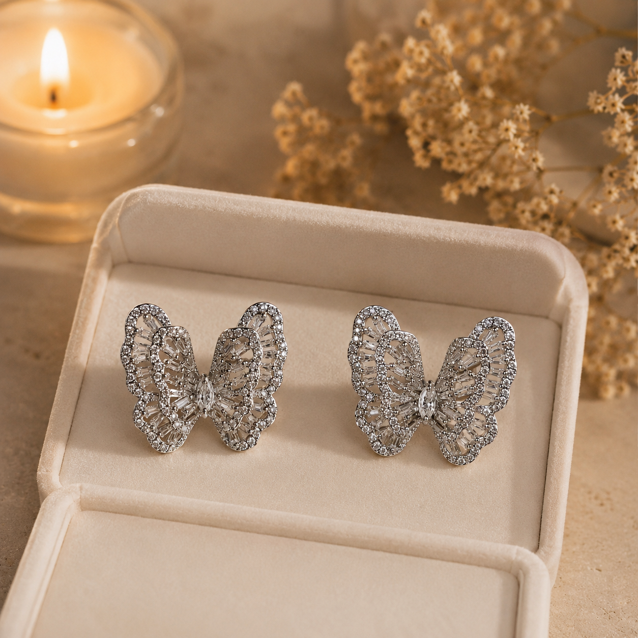 Silver crystal butterfly earrings displayed inside a premium ivory pearl-white velvet jewelry box with warm candlelight and dried floral accents in the background