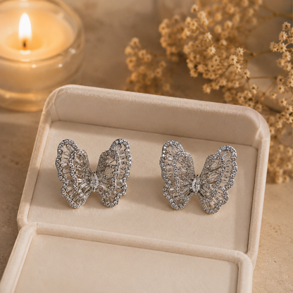 Silver crystal butterfly earrings displayed inside a premium ivory pearl-white velvet jewelry box with warm candlelight and dried floral accents in the background
