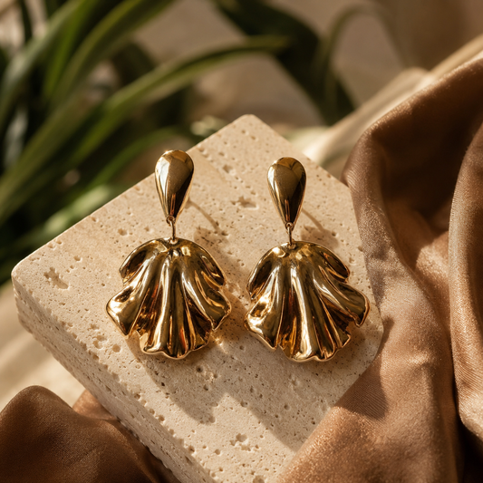 Artisan gold shell drop earrings displayed on cream travertine stone with mocha silk accents and warm natural light.