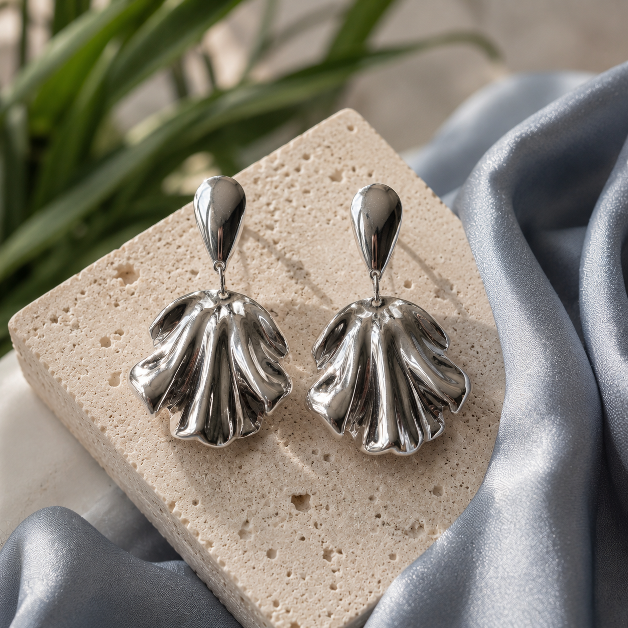 Sculptural silver shell drop earrings displayed on cream travertine stone with soft blue-grey silk accents and blurred botanical background.
