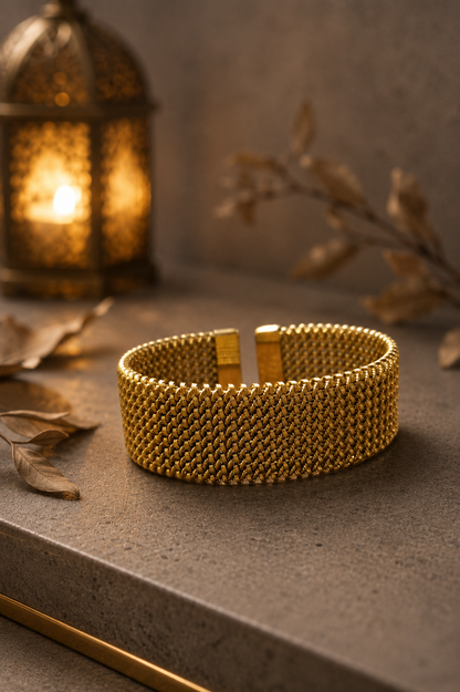 Gold-tone woven mesh cuff bracelet displayed on a luxury concrete surface with warm lantern lighting and dried botanical accents.