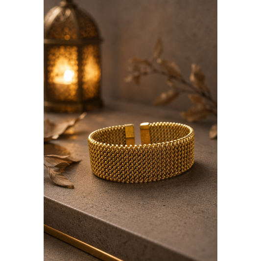 Gold-tone woven mesh cuff bracelet displayed on a luxury concrete surface with warm lantern lighting and dried botanical accents.