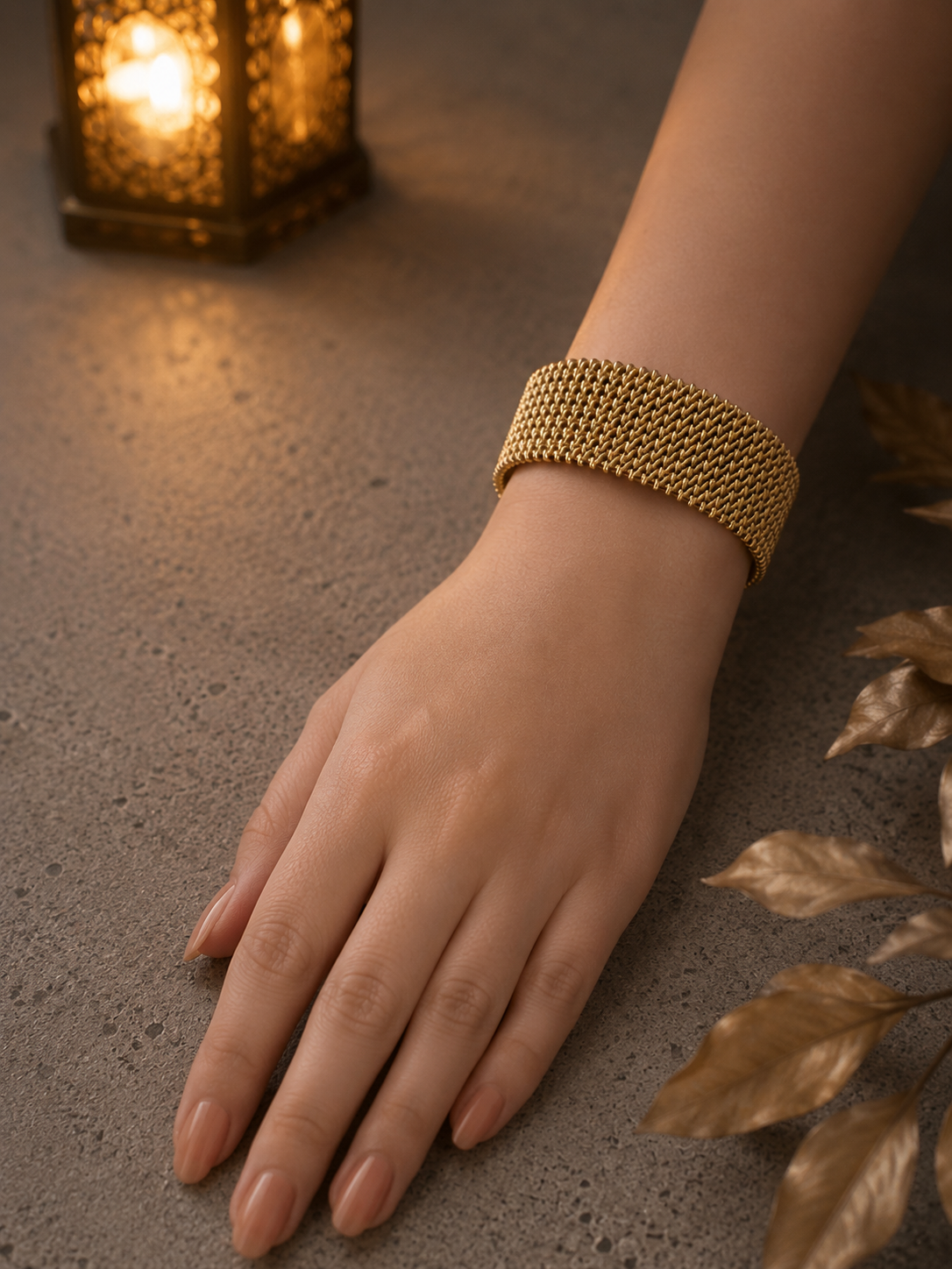 Woman wearing a flexible gold-tone woven mesh cuff bracelet on the wrist against a luxury concrete tabletop with warm ambient lantern lighting.