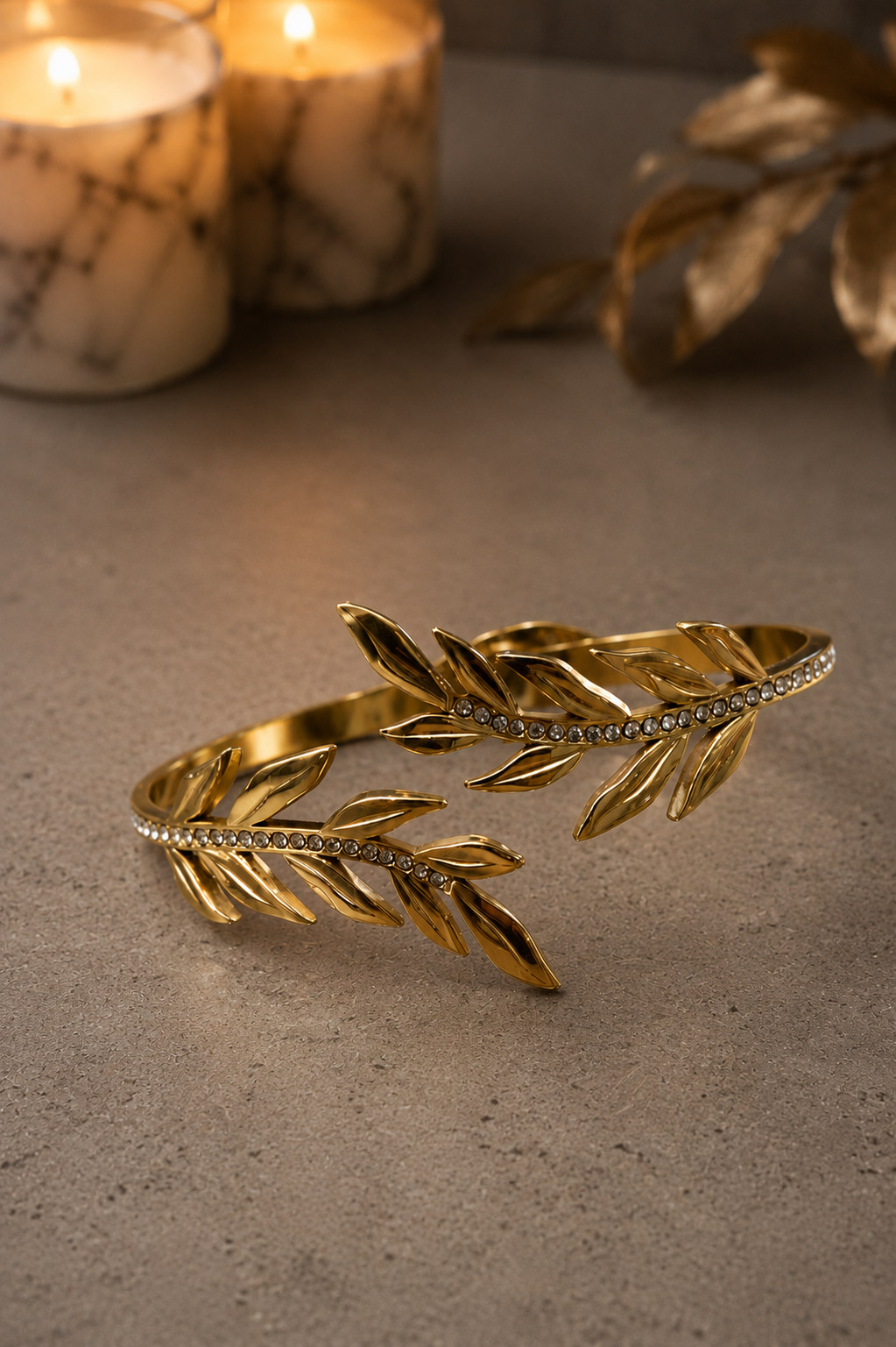 Gold-tone laurel leaf open cuff bracelet with crystal accents displayed on a luxury concrete surface with marble candles and warm ambient lighting.