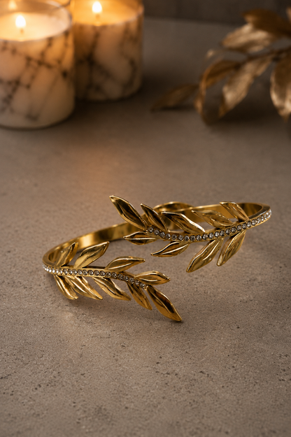 Gold-tone laurel leaf open cuff bracelet with crystal accents displayed on a luxury concrete surface with marble candles and warm ambient lighting.