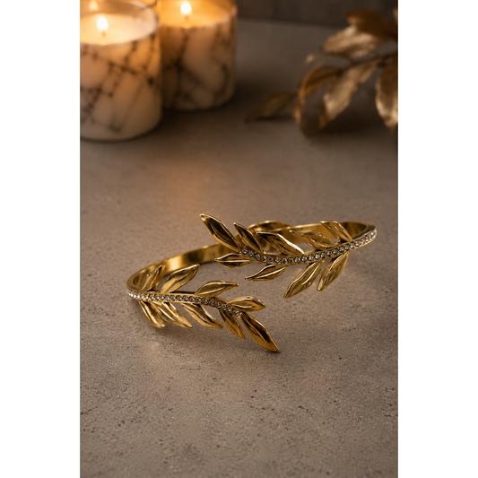 Gold-tone laurel leaf open cuff bracelet with crystal accents displayed on a luxury concrete surface with marble candles and warm ambient lighting.