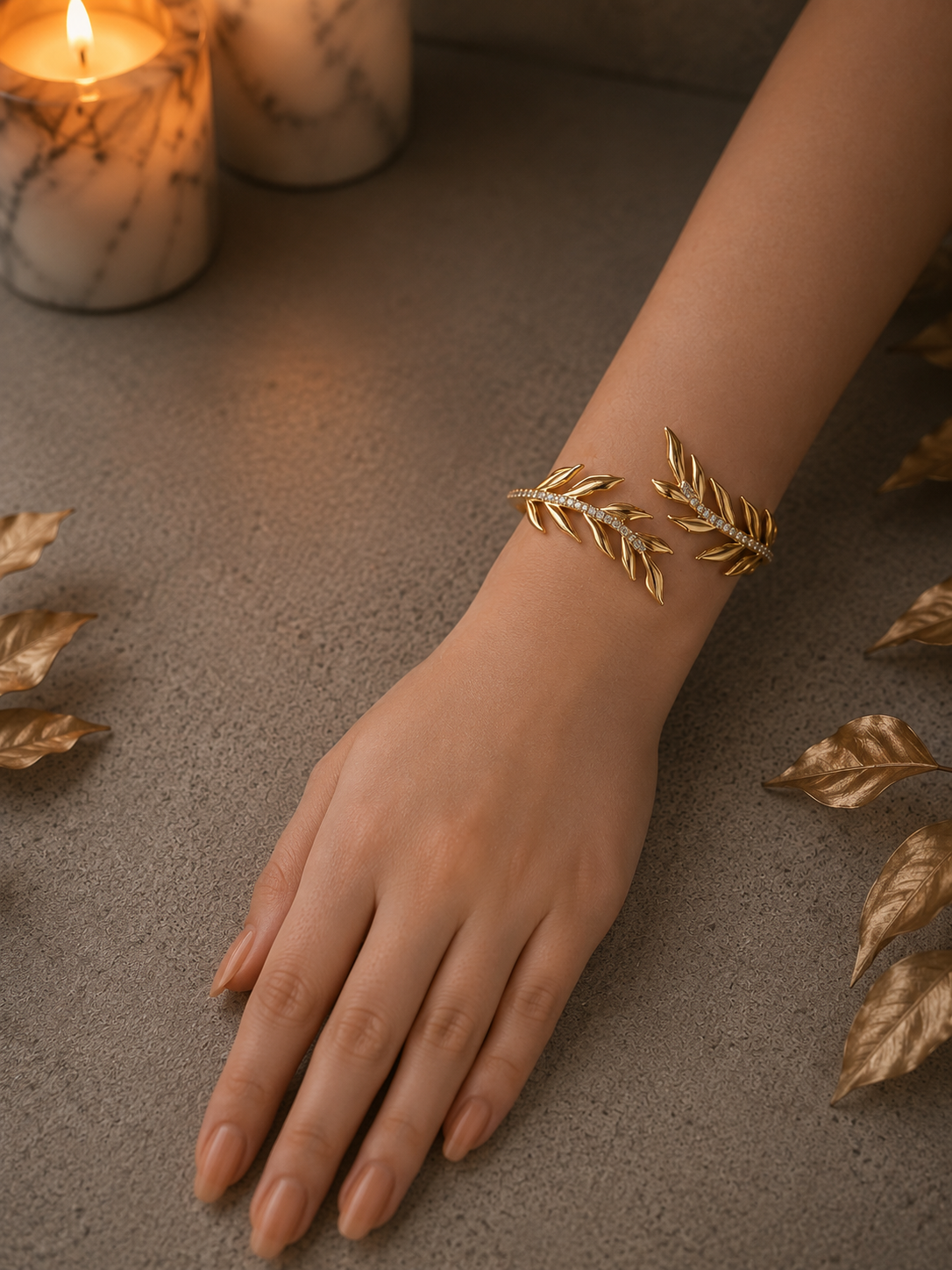 Woman wearing a gold laurel leaf crystal cuff bracelet on the wrist with a luxury concrete background, marble candles, and decorative botanical styling.