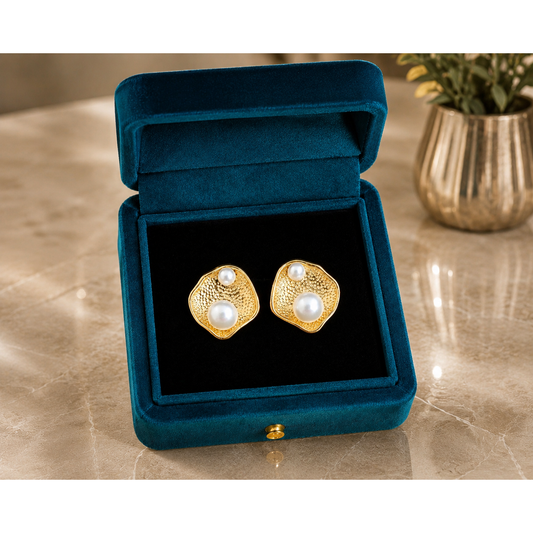Organic gold pearl stud earrings displayed inside a premium teal velvet jewelry box on a reflective marble surface with a blurred decorative plant in the background.