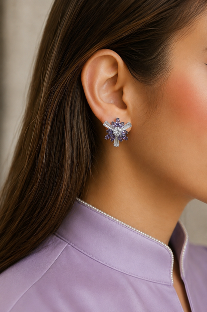 Model wearing silver-tone floral stud earrings with lilac marquise crystals and baguette-cut stones, styled with a soft lilac satin shirt featuring pearl-trimmed neckline.