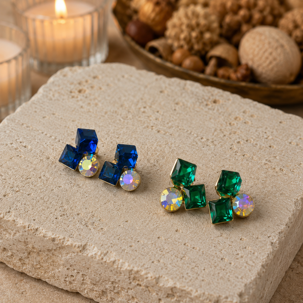 Pair of blue and green gemstone earrings on a stone surface with a candle and decorative items in the background.