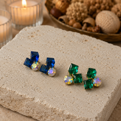 Pair of blue and green gemstone earrings on a stone surface with a candle and decorative items in the background.