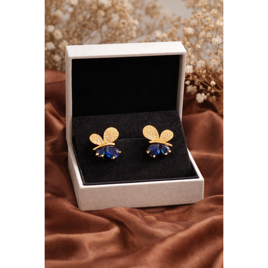 Butterfly stud earrings displayed in jewelry box on brown silk cloth with blurred dry flowers background in gold and blue stone variant